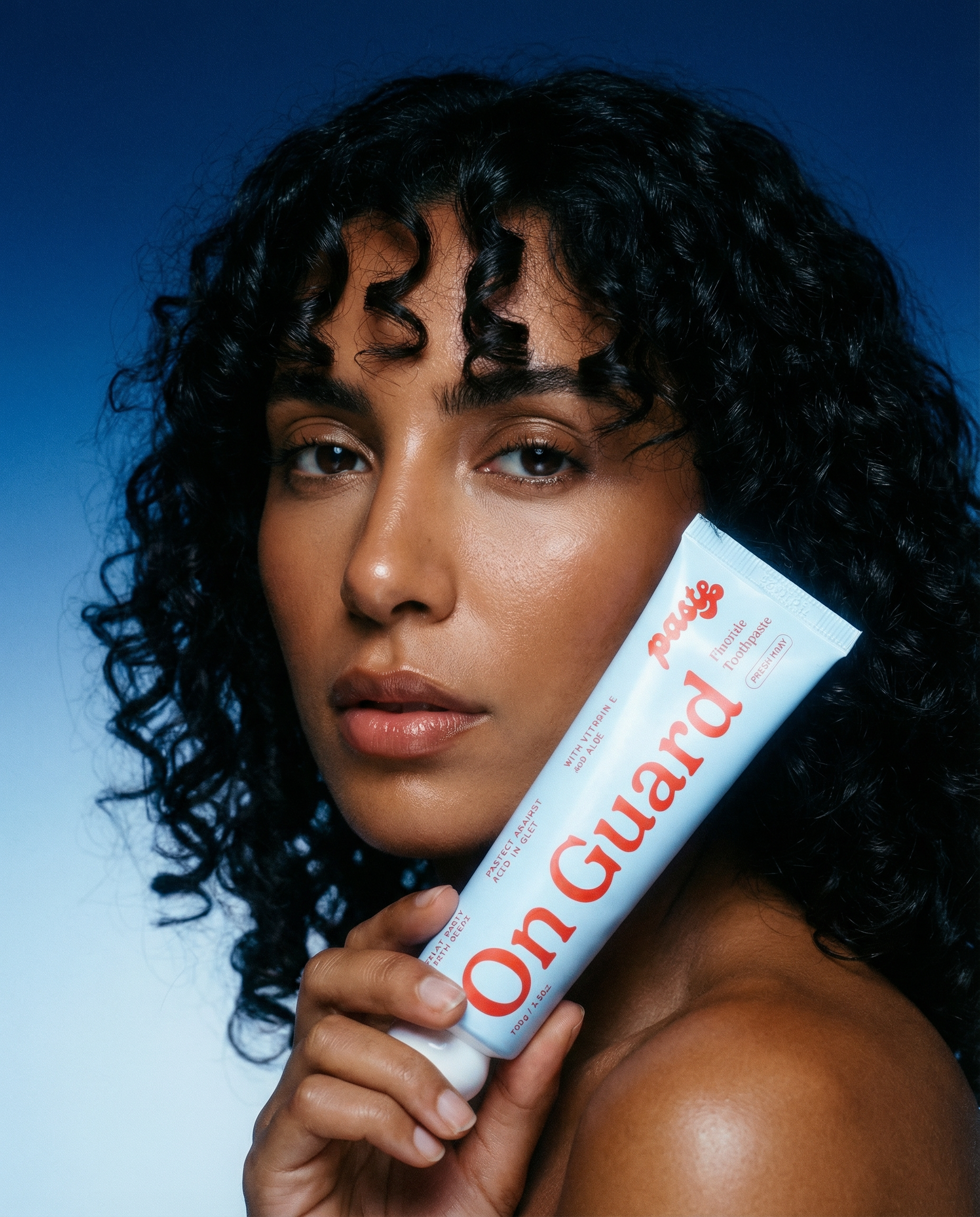 Woman with curly dark hair holds a tube of Paste On Guard fluoride toothpaste beside her face against a deep blue gradient background.