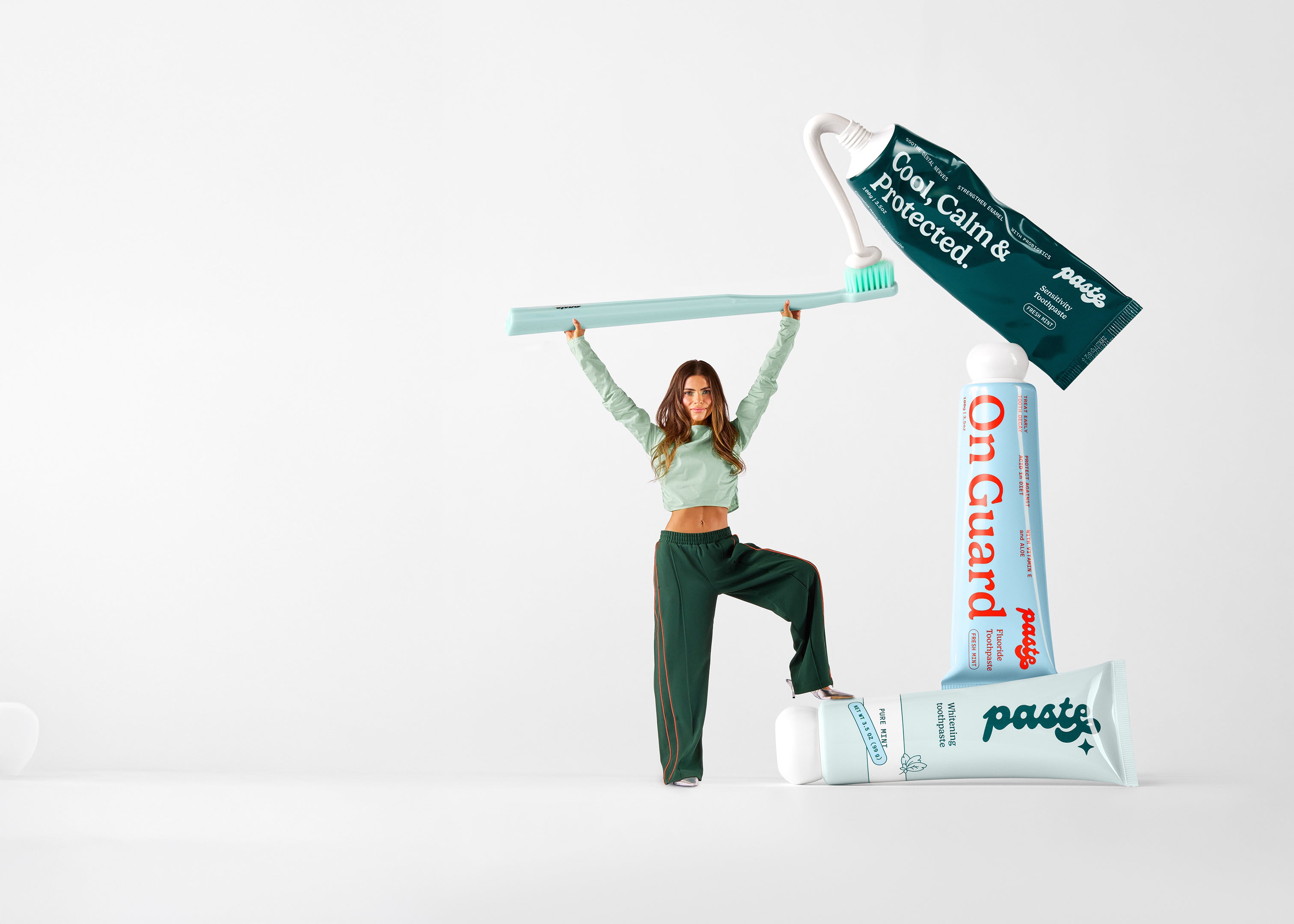 Woman holding a toothbrush over her head with giant life size sensitivity toothpaste, fluoride toothpaste, and whitening toothpaste next to her. White background. 