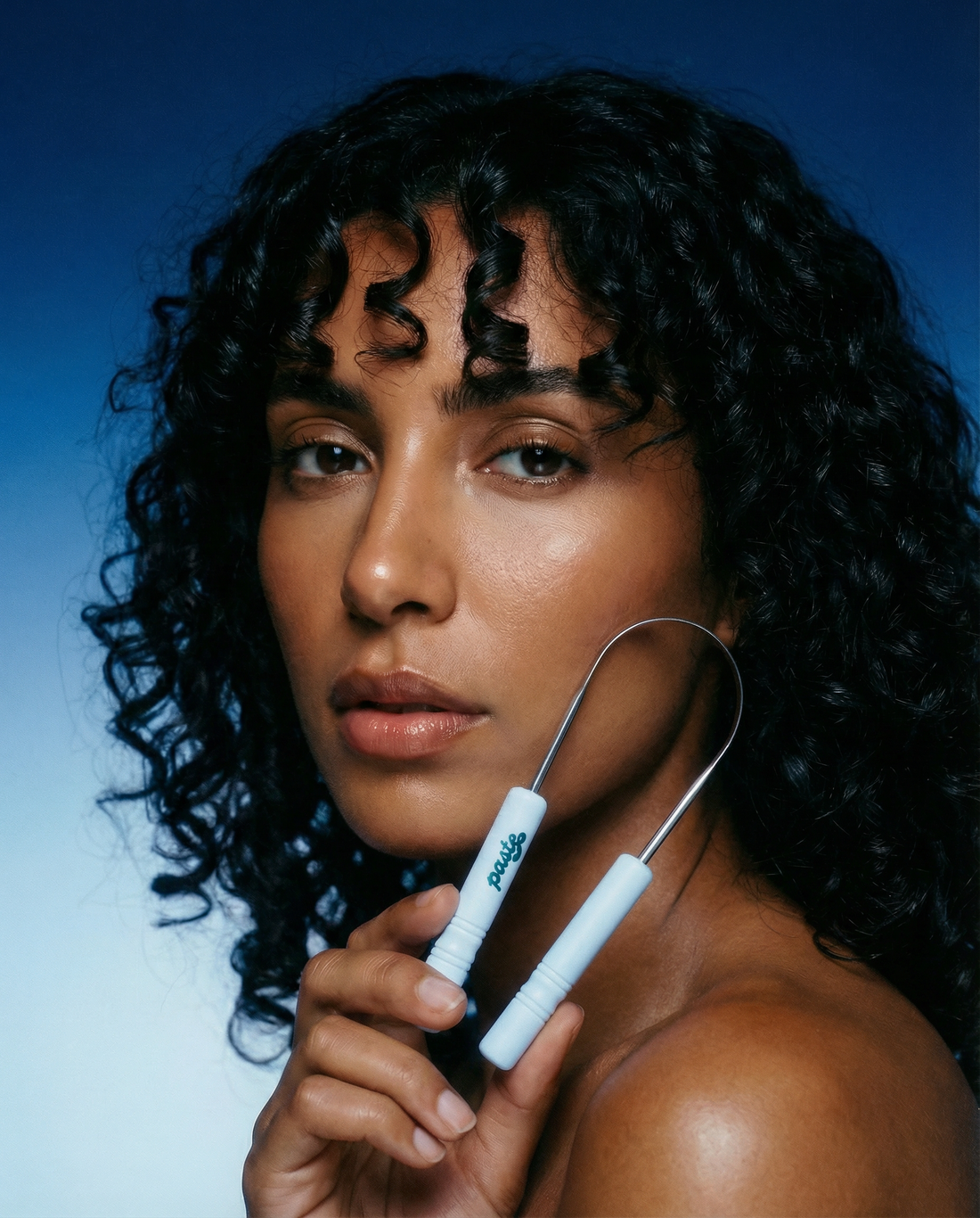Woman with curly dark hair holds a Paste tongue scraper with white handles and a stainless steel arch against her jawline, on a deep blue-to-light blue gradient background.