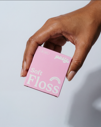 A hand with natural nails holds a pink box of Paste Soft Floss in Watermelon flavor (30 uses) against a light grey-blue background.